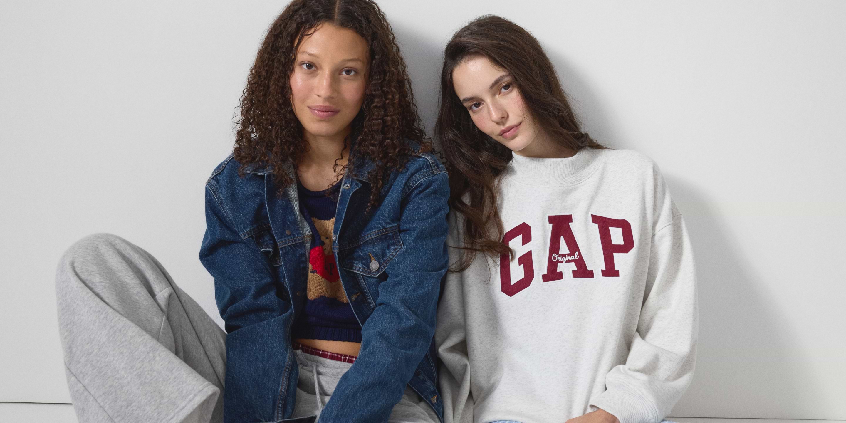 Two models sit against a white wall—one in a denim jacket and gray sweats, the other in a light gray GAP sweatshirt.