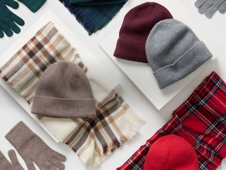 Flat lay of winter accessories: knit beanies, plaid scarves, and gloves in burgundy, gray, taupe, red, and green tones on a white background.