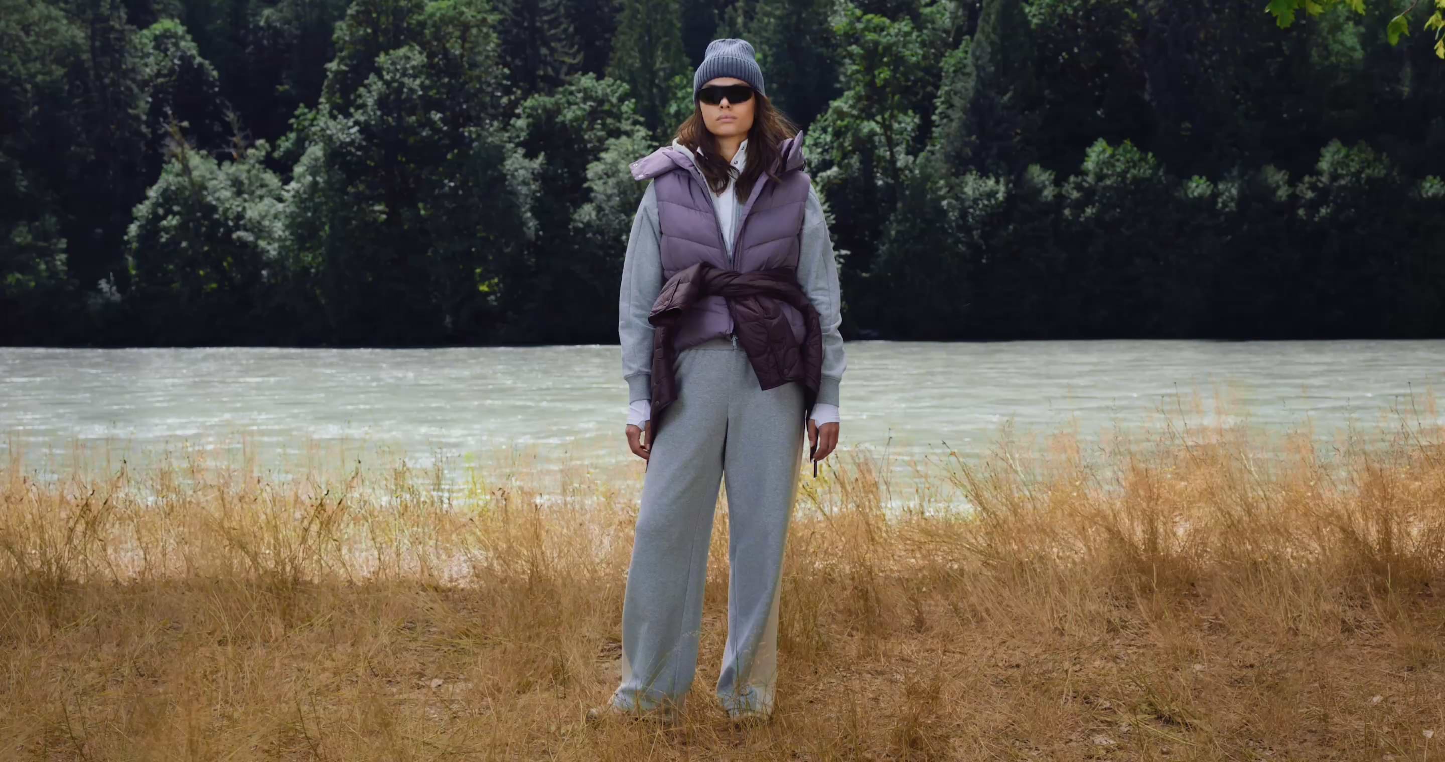 Person in a beanie, sunglasses, purple puffer vest and gray sweats stands on dry grass by a river, with dense forest in the background.