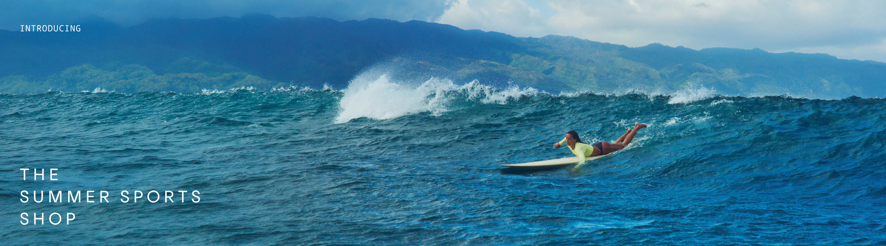 Surfer paddling on a blue ocean wave with mountains in the background.