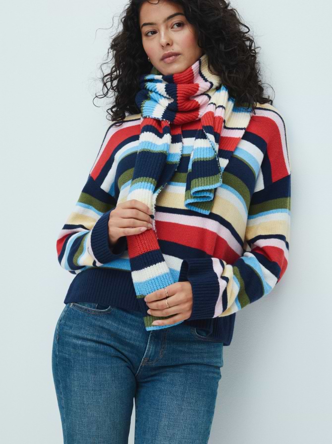 Person with curly hair wearing a multicolored striped knit sweater and matching scarf with blue jeans against a plain light background.
