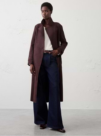 Person in a long brown coat over a white top and wide-leg dark jeans, standing in a studio.