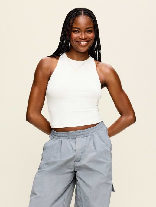 Person smiling with hands behind back, wearing a white sleeveless crop top and gray pants on a plain background.