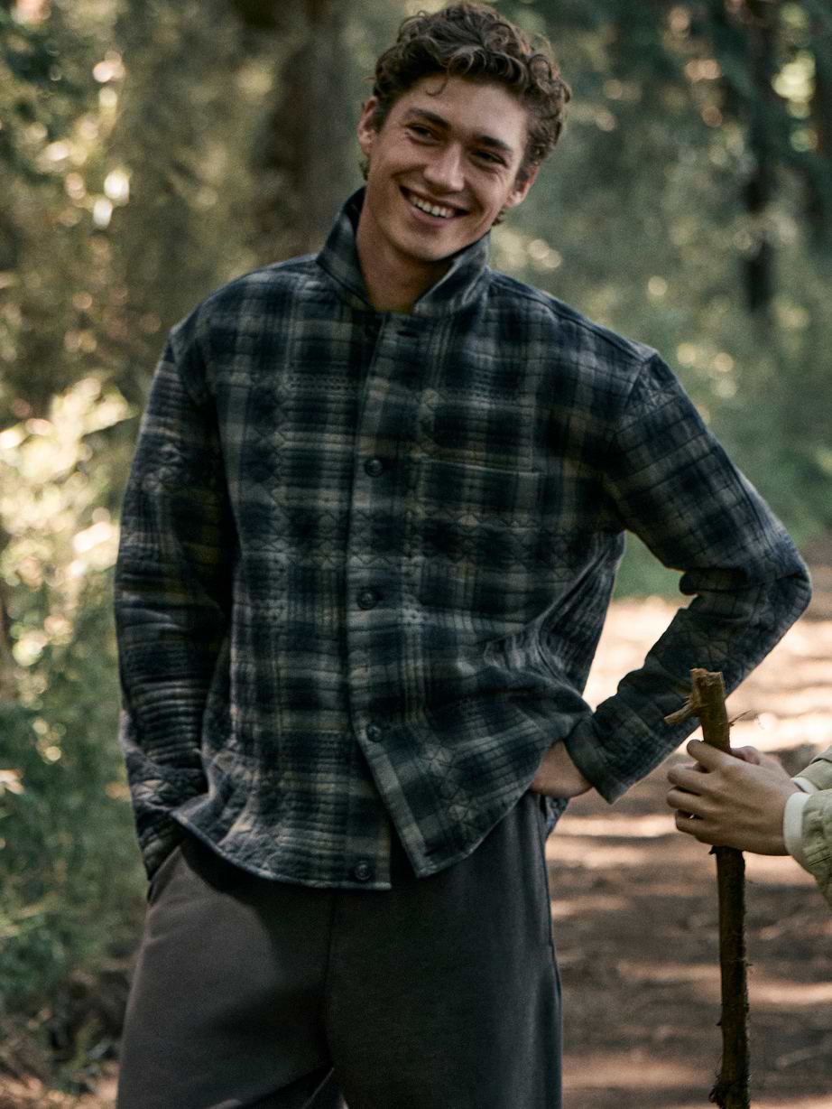 Smiling person in a gray plaid jacket on a forest trail, with another person’s hand holding a walking stick beside them.