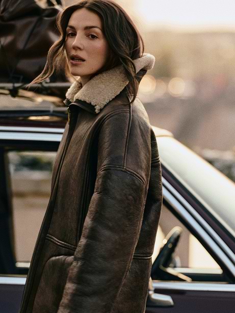 Person in a brown shearling coat stands beside a classic car with roof luggage at dusk.