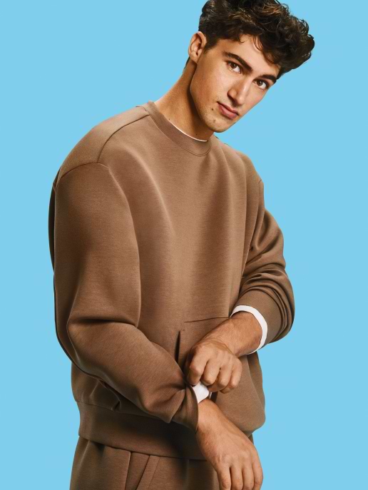 Person in a brown sweatshirt rolling up a sleeve against a light blue background.