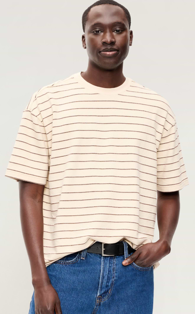 Person wearing a beige short‑sleeve T‑shirt with thin horizontal stripes and blue jeans, standing against a plain light background.