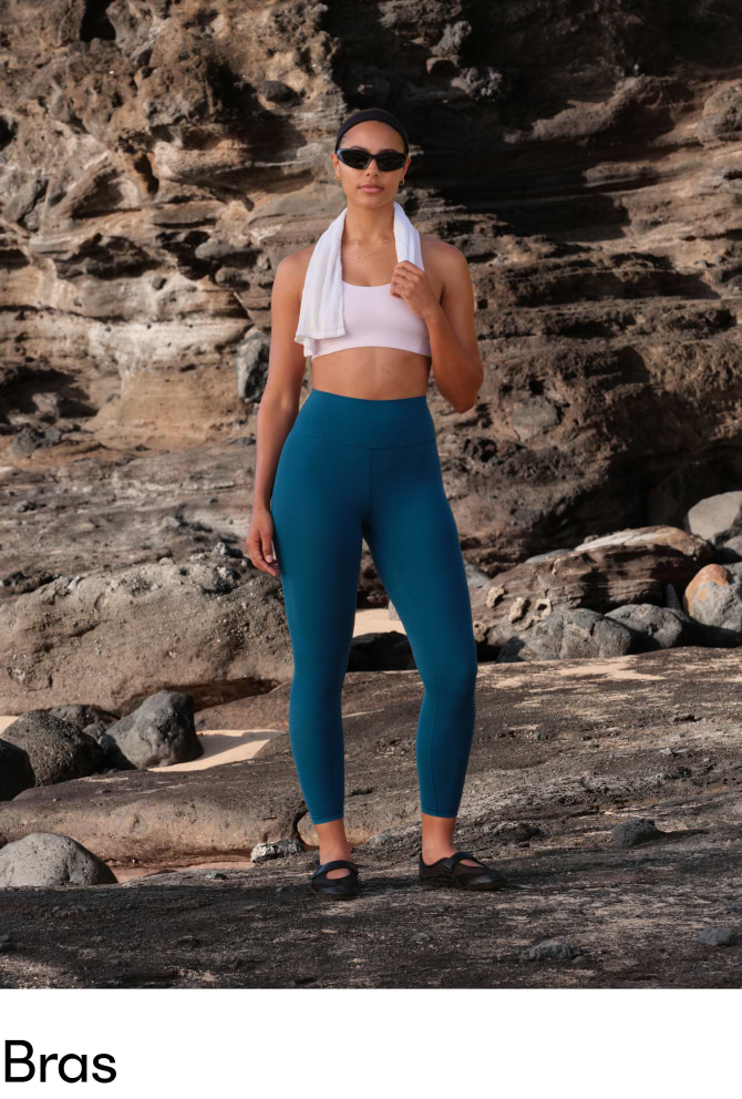 Person standing on a rocky shore wearing a white sports bra, teal leggings, and a towel around their neck.