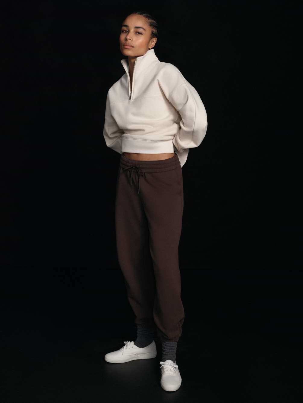 Person in a cream half‑zip cropped sweatshirt and brown joggers with white sneakers, standing against a black background.