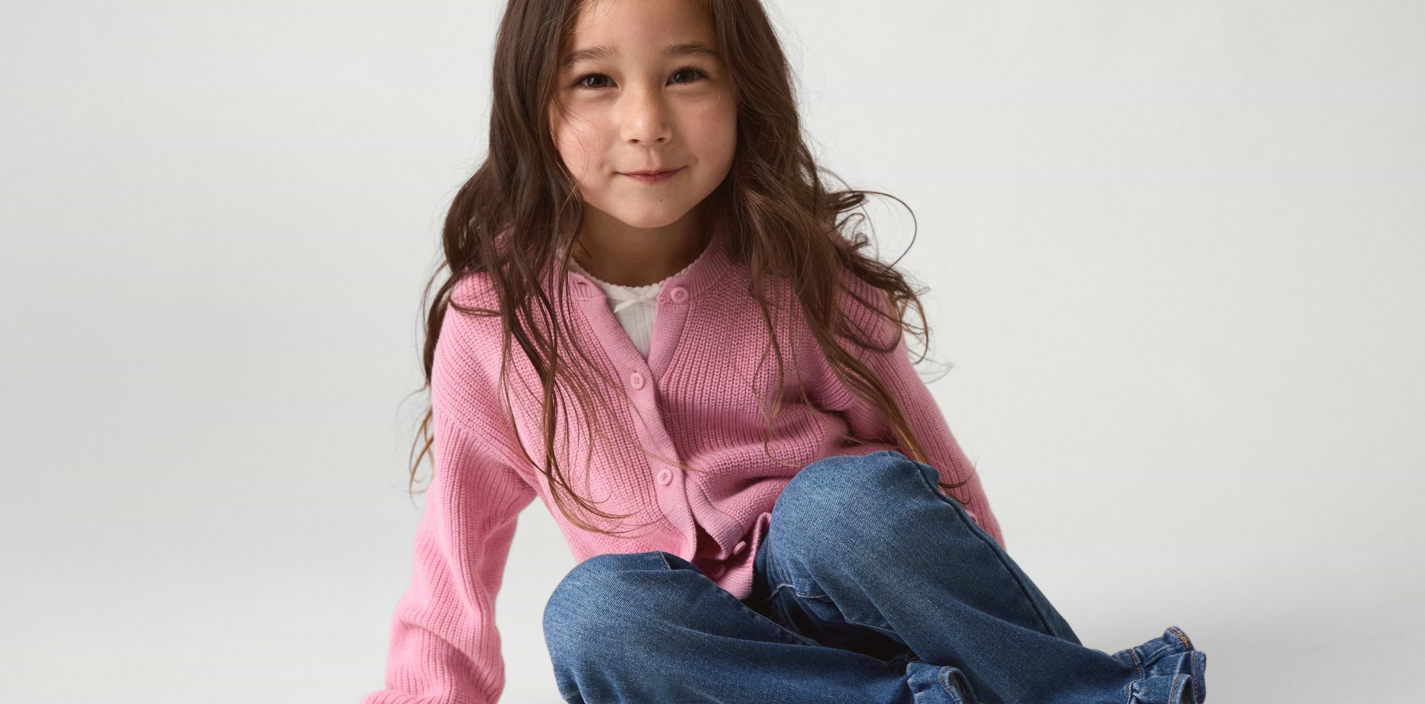 Smiling child in a pink cardigan and blue jeans sitting on the floor against a plain background.