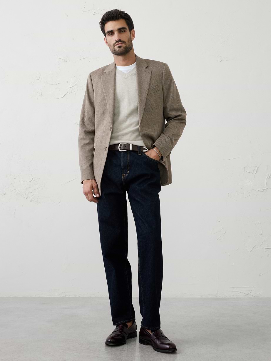 Man standing against a plain wall wearing a beige blazer over a light sweater, dark jeans, and burgundy loafers.