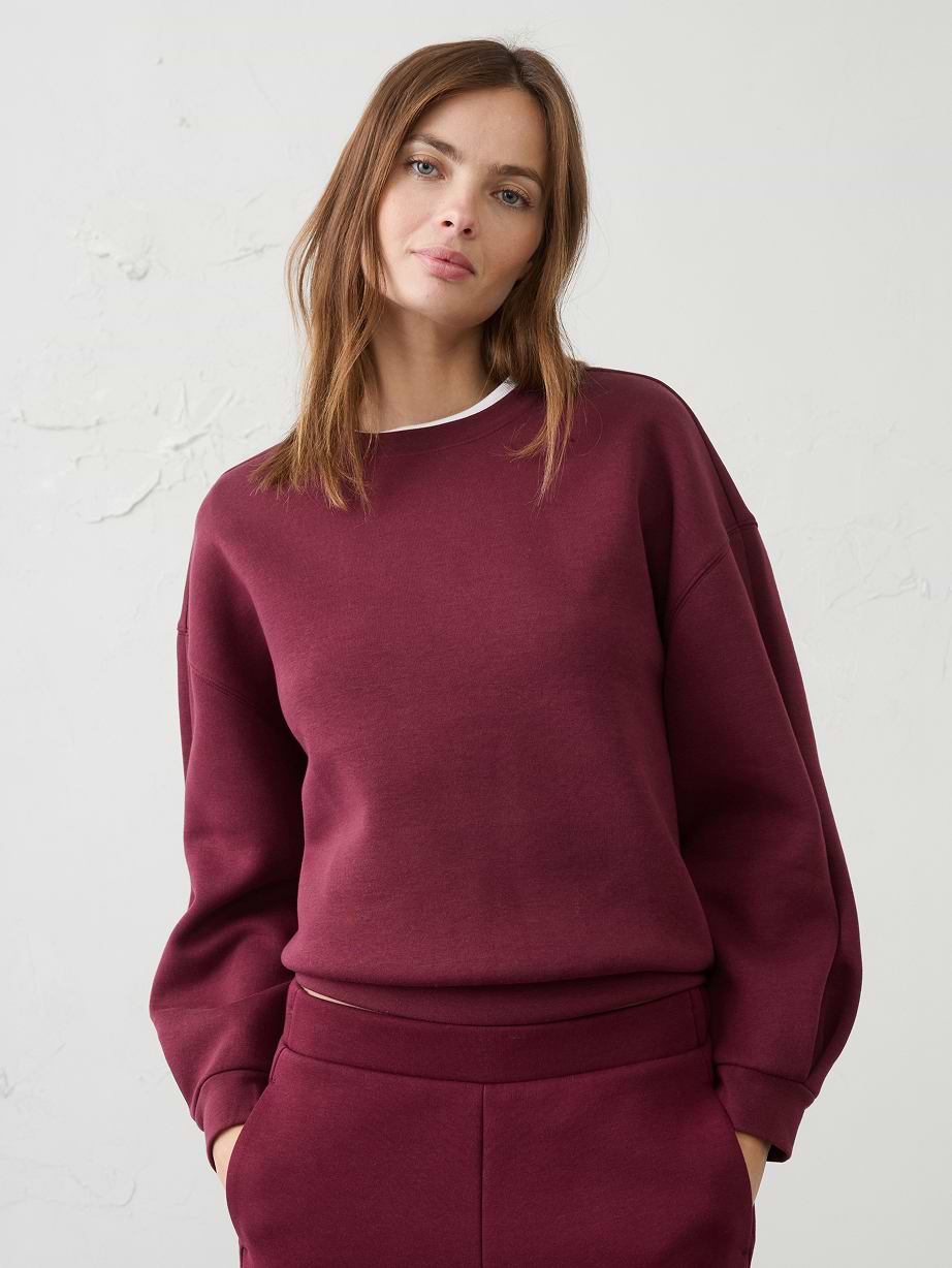 Person in a burgundy sweatshirt and matching joggers against a plain light wall.