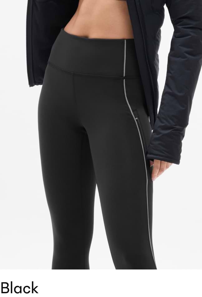 Close-up of a person wearing black high-waisted leggings with grey piping along the side, partially covered by a black jacket.