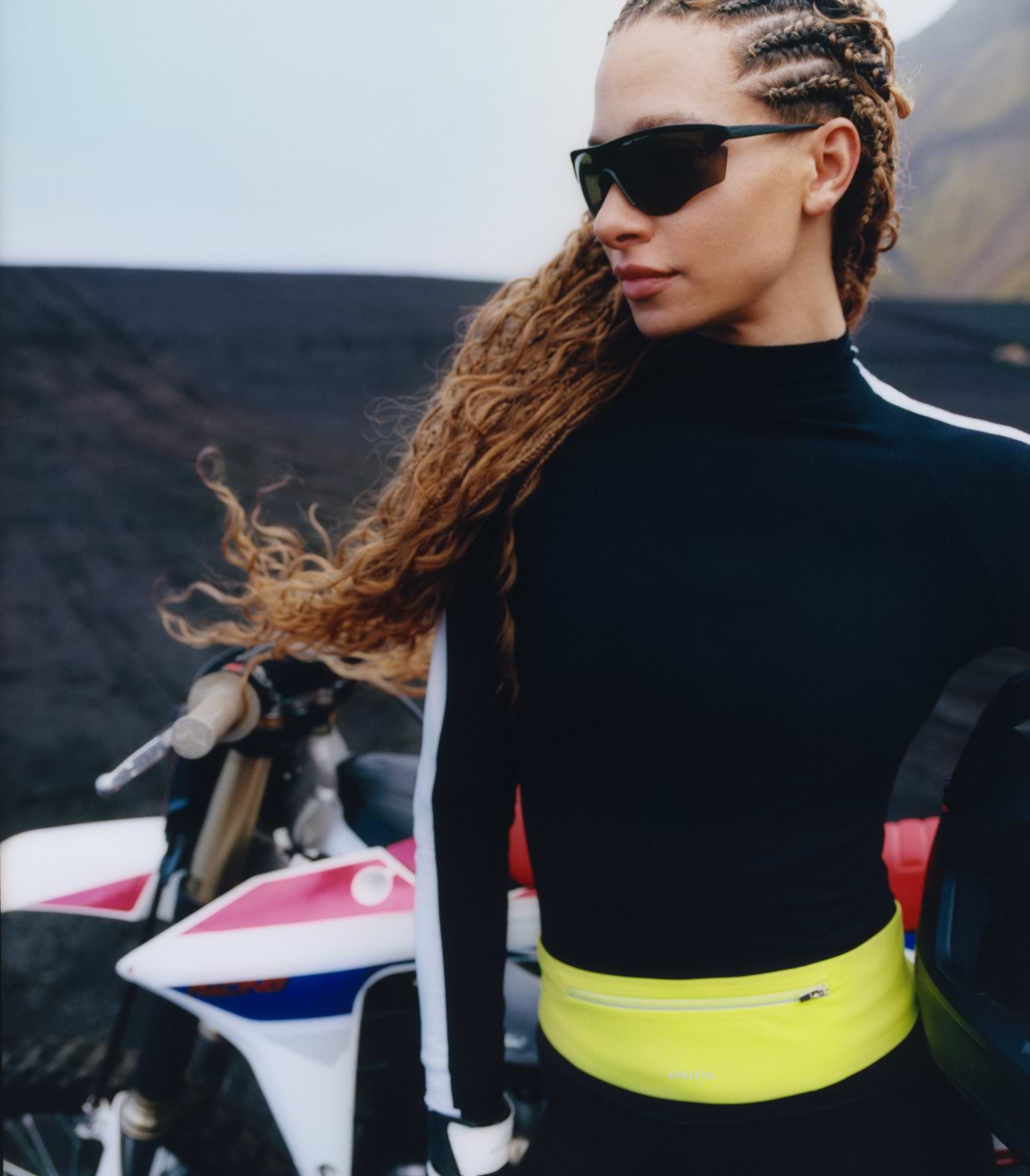 Person with braided hair and sunglasses stands beside a white dirt bike on dark volcanic terrain, wearing a neon yellow waist pack.