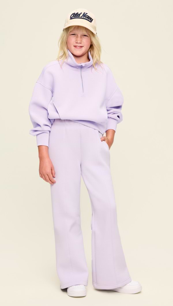 Child in a lavender quarter-zip sweatshirt and matching wide-leg pants, white sneakers, and a beige cap reading “Old Navy,” standing against a plain light background.