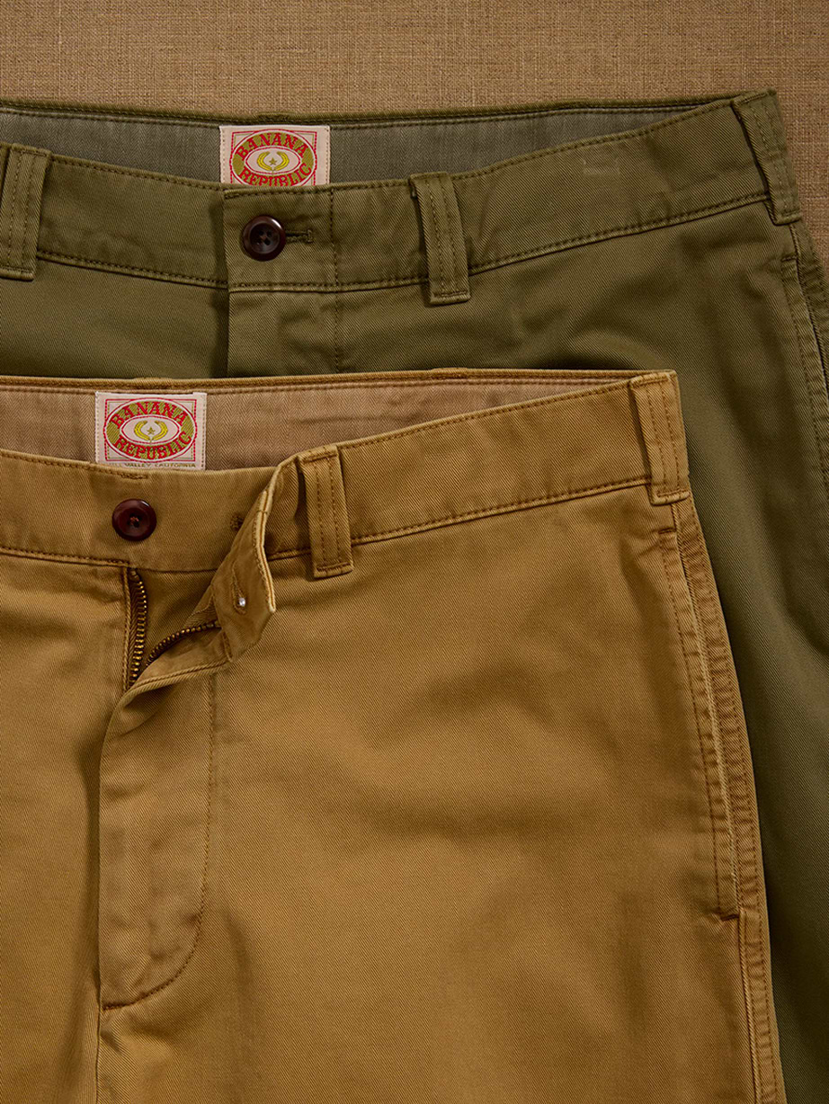 Two pairs of chinos, olive and tan, overlapped on beige fabric with the tan fly open.