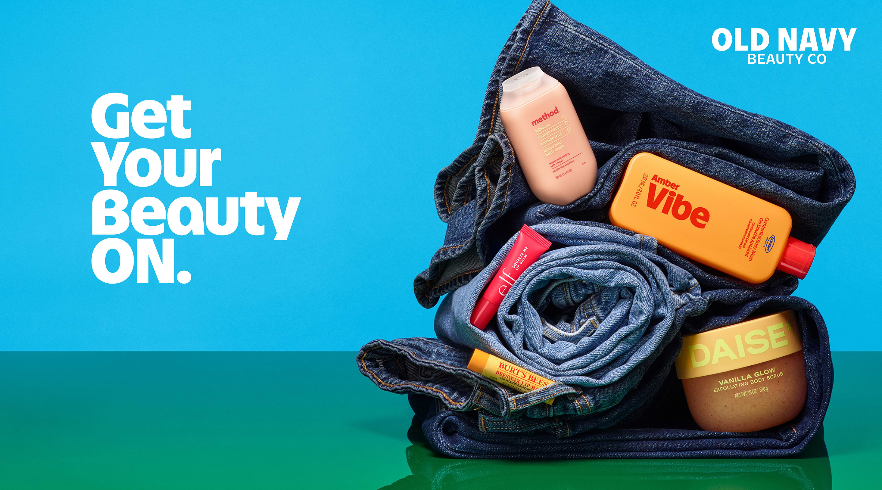 Rolled blue jeans stacked on a green surface with beauty items tucked in—orange Amber Vibe bottle, pink Method wash, e.l.f. gloss, Burt’s Bees balm, and DAISE scrub—under “OLD NAVY BEAUTY CO” on a blue background.