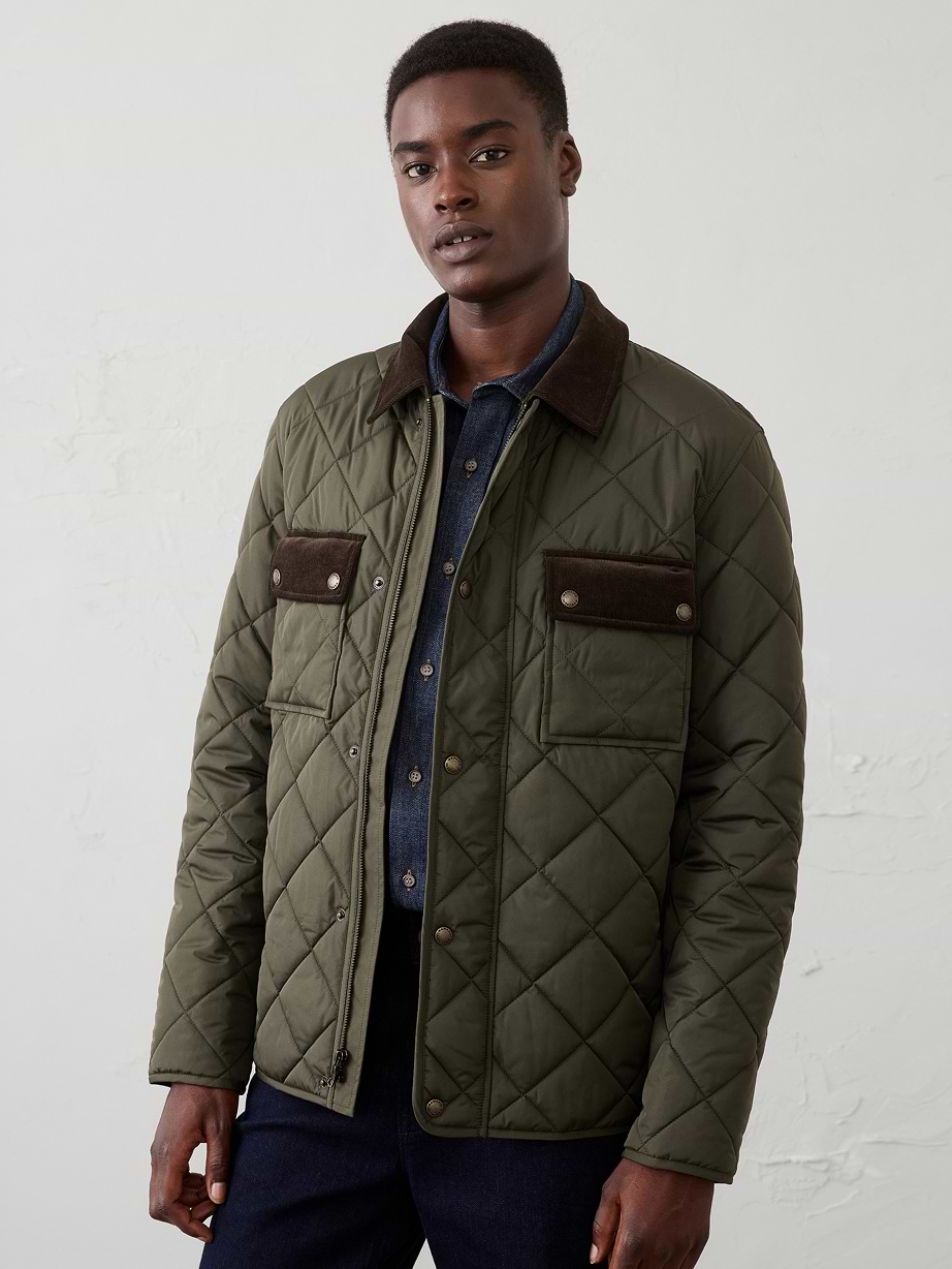 Person wearing an olive green diamond‑quilted jacket with dark brown collar and flapped chest pockets, standing against a plain light background.