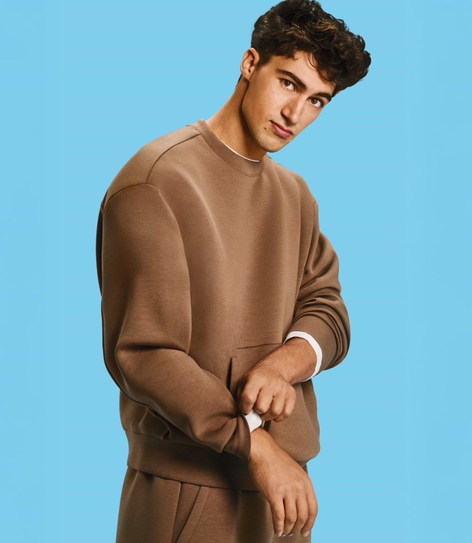 Person in a brown sweatshirt and matching pants rolling up a sleeve against a blue background.