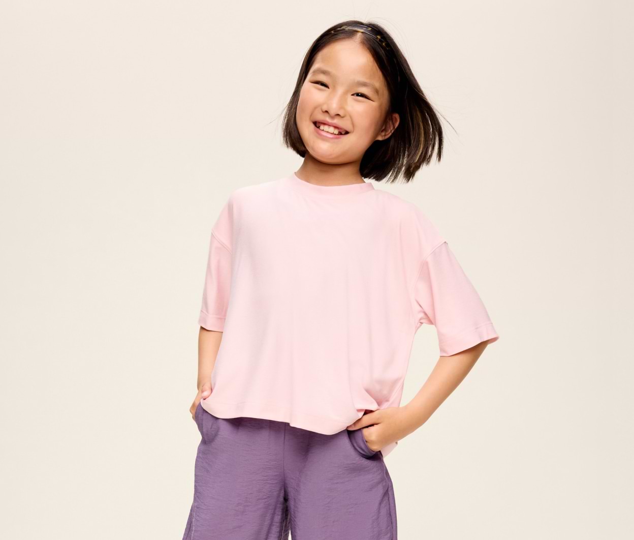 Smiling child in a light pink T-shirt and purple shorts, hands in pockets, against a plain background.