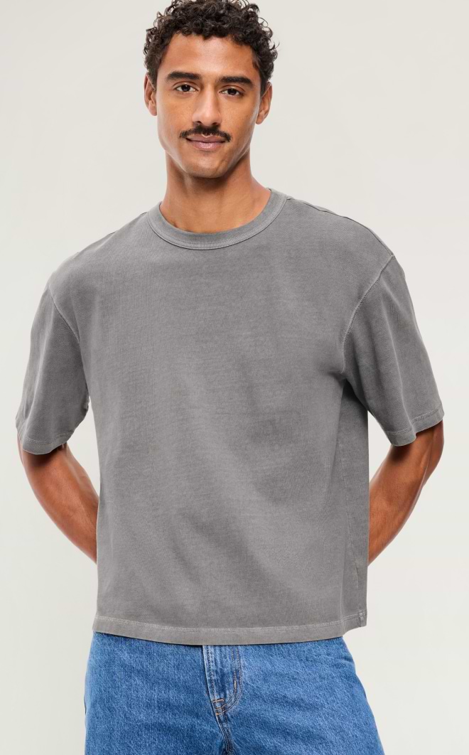 Person wearing a gray short-sleeve T-shirt and blue jeans, standing with hands behind their back against a plain background.