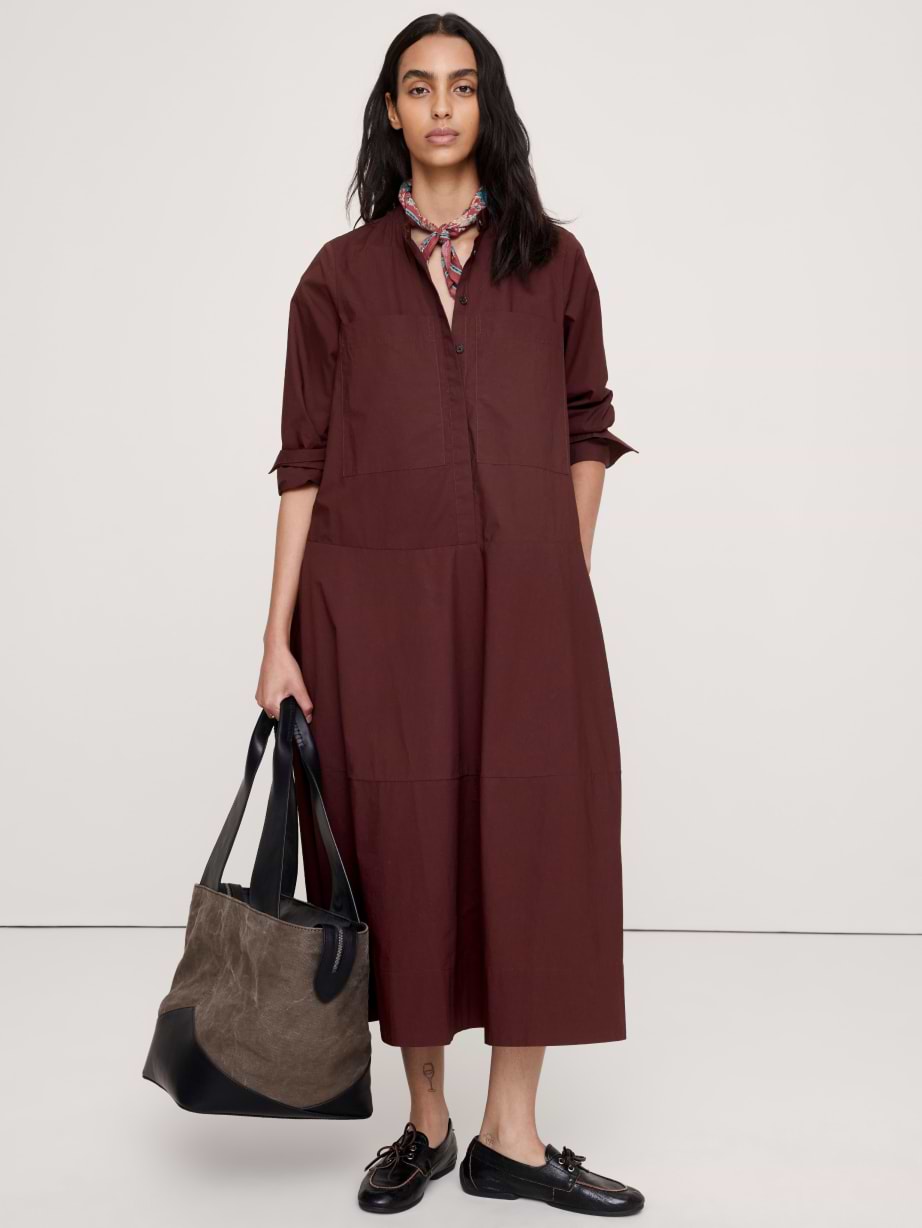 Person in a long maroon shirt dress with rolled sleeves and a patterned neck scarf, holding a two‑tone tote, black loafers, standing against a plain backdrop.