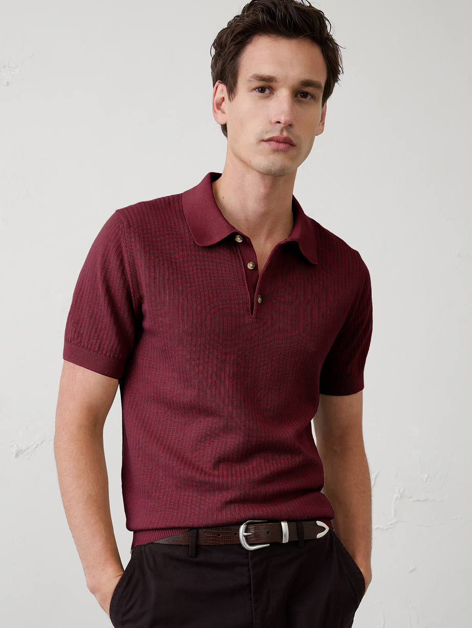 Person wearing a burgundy short-sleeve knit polo and dark pants against a plain wall.