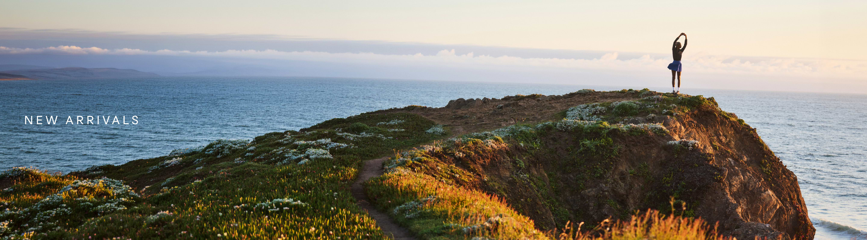 Woman with arms raised in a stretching position on a grassy sea cliff at sunset, overlooking the ocean; text reads “New Arrivals.”