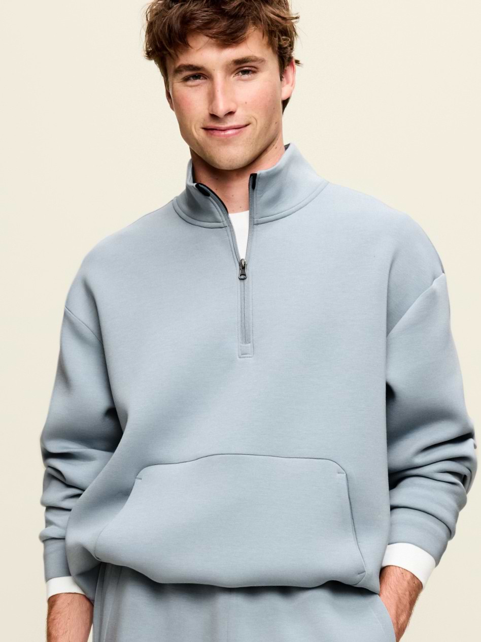 Model wearing a light blue quarter‑zip sweatshirt with stand collar and front kangaroo pocket, against a plain background.