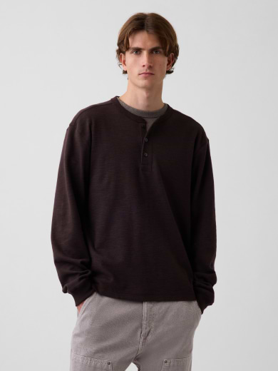 Une personne porte un haut henley foncé à manches longues par-dessus un t-shirt gris et un pantalon gris pâle, debout devant un arrière-plan uni pâle, les mains dans les poches.