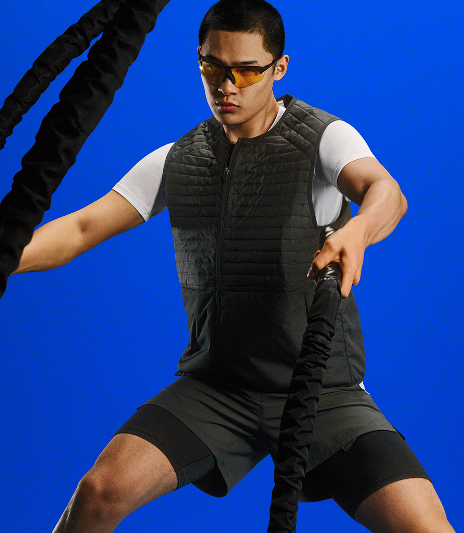 Person exercising with battle ropes, wearing sport sunglasses, a white tee, and a black padded vest against a bright blue background.