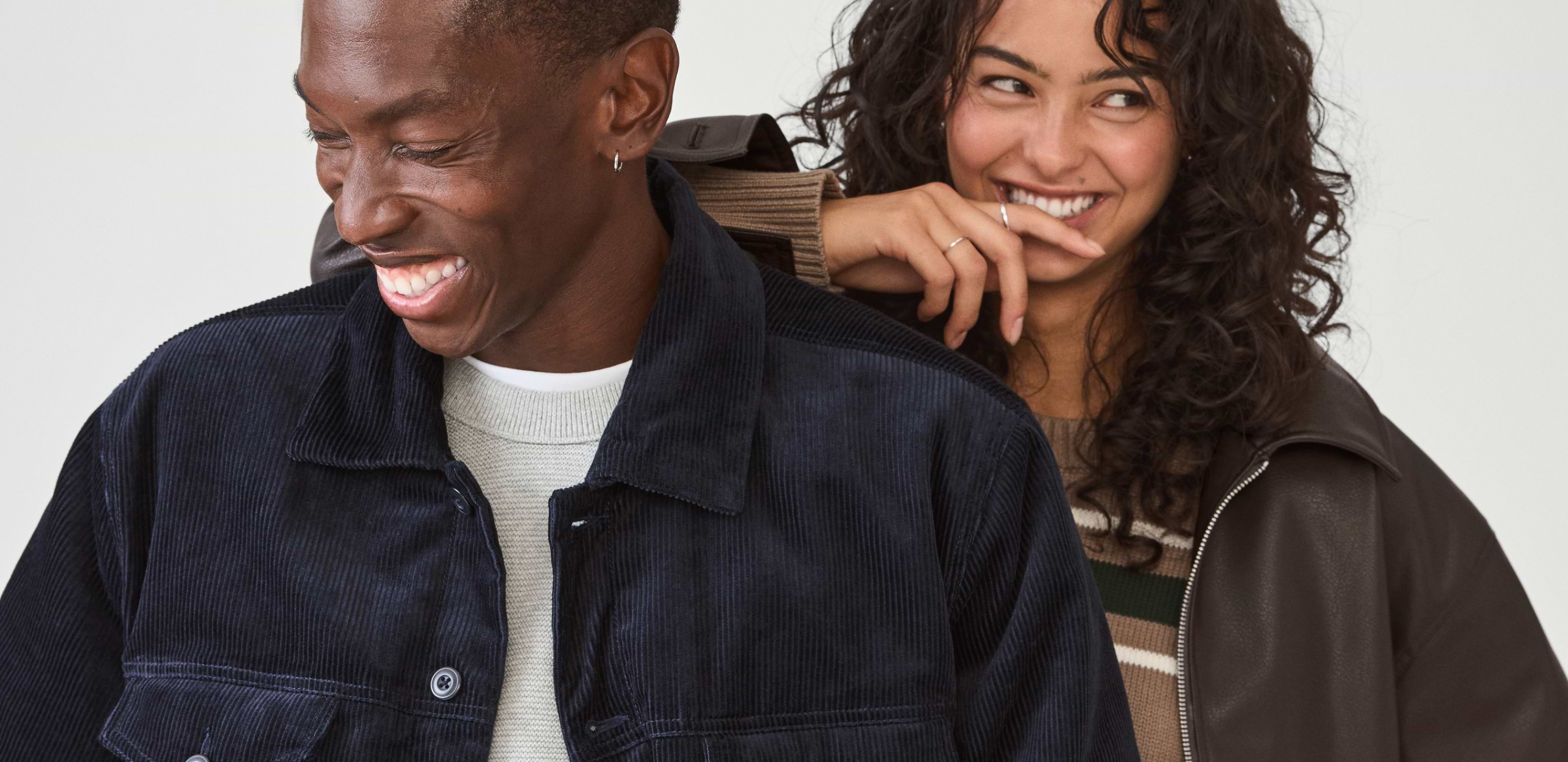 Two people smiling together—one in a navy corduroy jacket in front, the other with curly hair in a brown jacket and striped sweater behind.