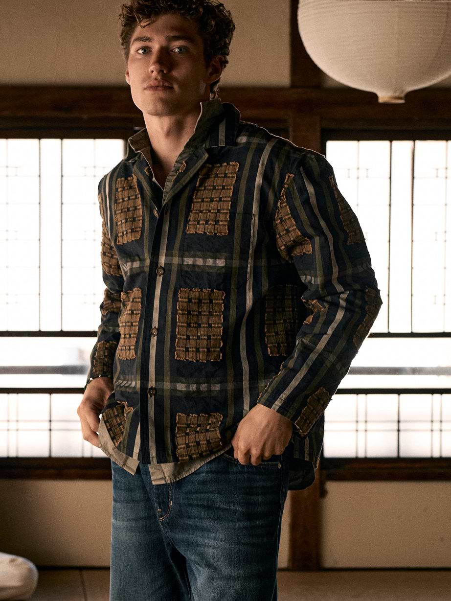 Person wearing a navy striped button-up with tan patchwork and blue jeans, standing in a softly lit room with paper-paneled windows.