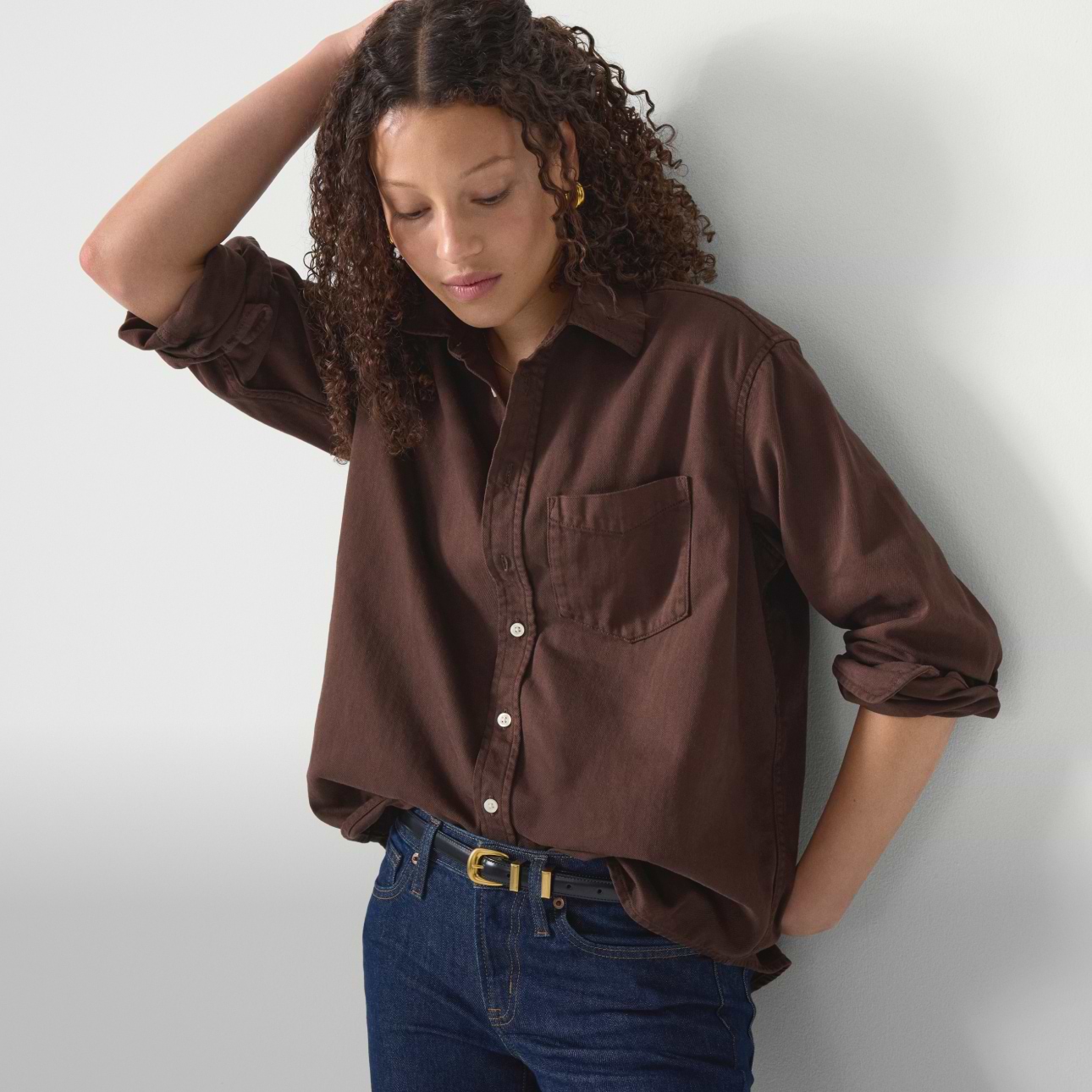 Person with curly hair wearing a brown button-up shirt and blue jeans, hand on head, looking down.