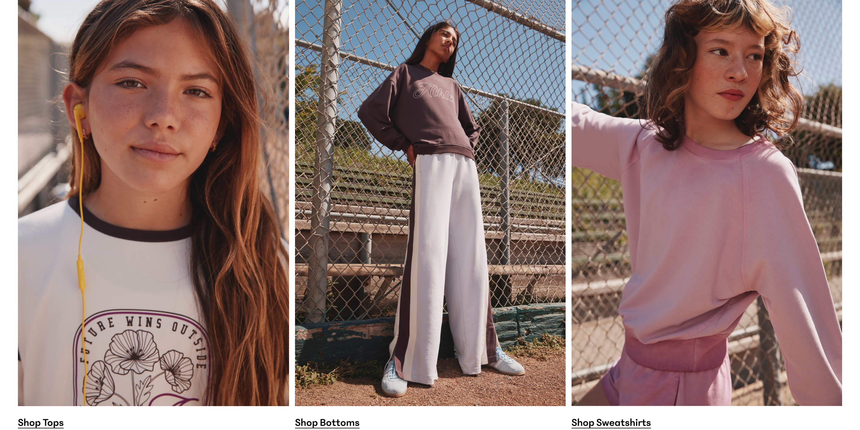 Three young girls by a chain-link fence modeling casual wear: white graphic top with earbuds, mauve sweatshirt with wide-leg pants, and pink sweatshirt with shorts.