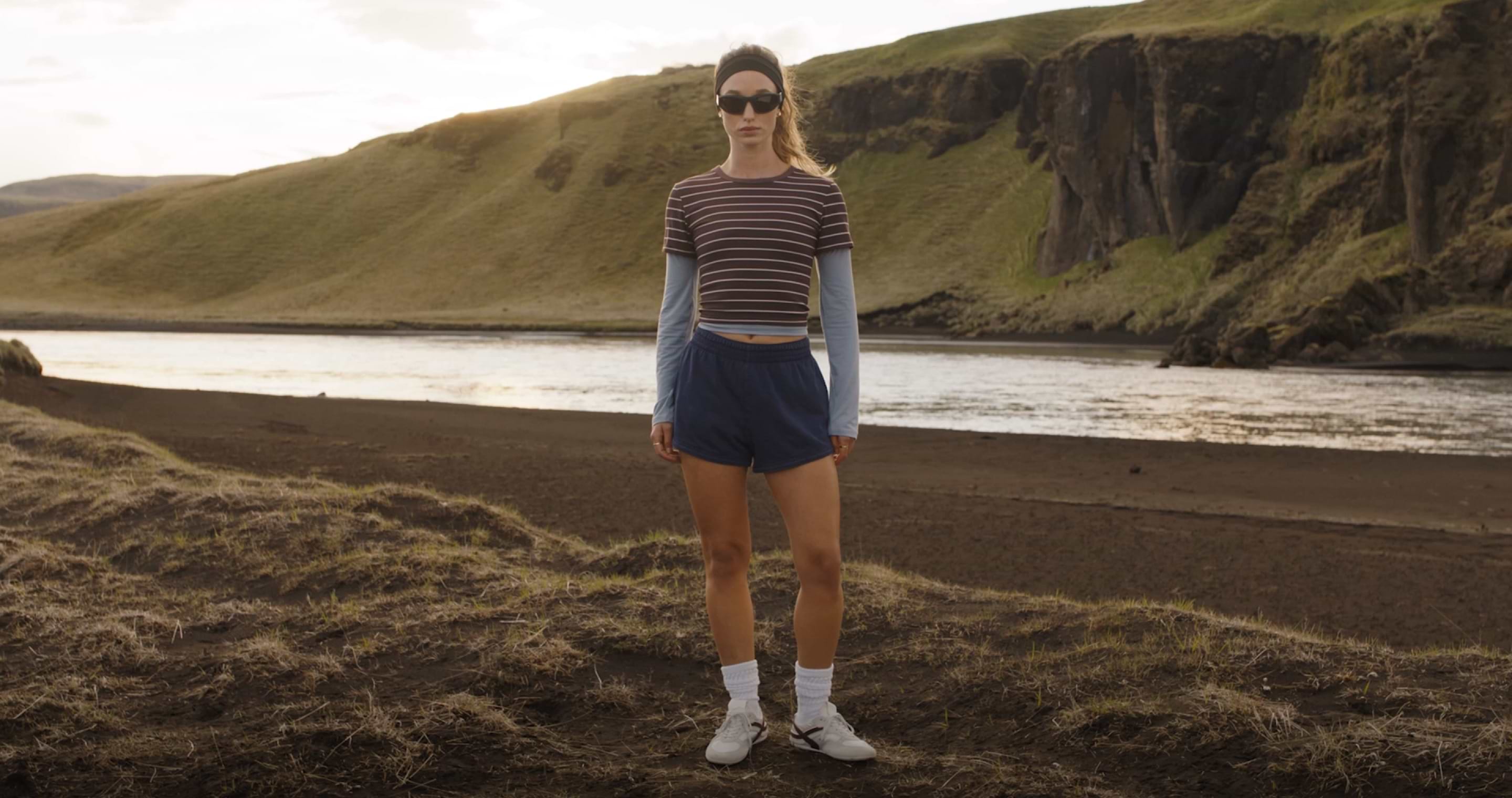 Woman in a striped crop top, blue shorts, and sneakers stands on a dark riverbank with green hills and cliffs behind.