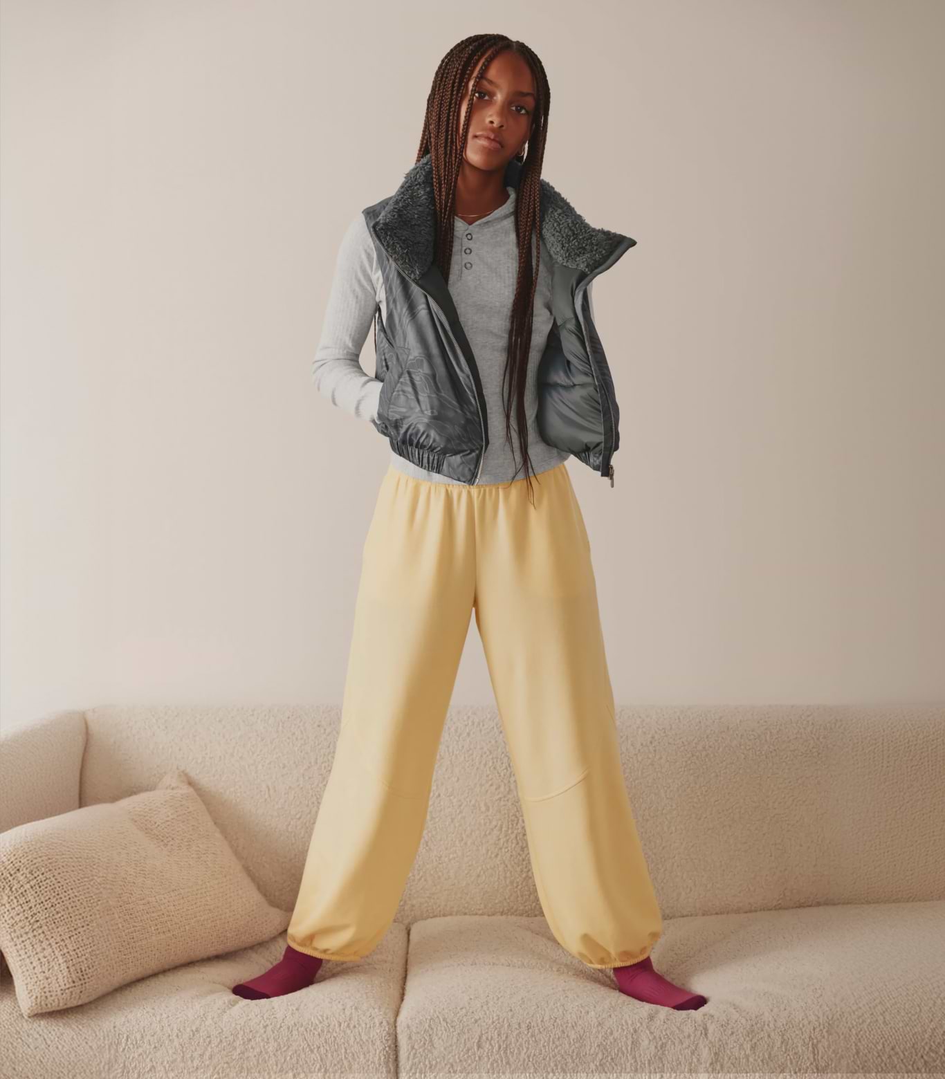 Girl with long braids stands on a beige sofa, wearing a gray henley, dark gray puffer vest, yellow joggers, and maroon socks.