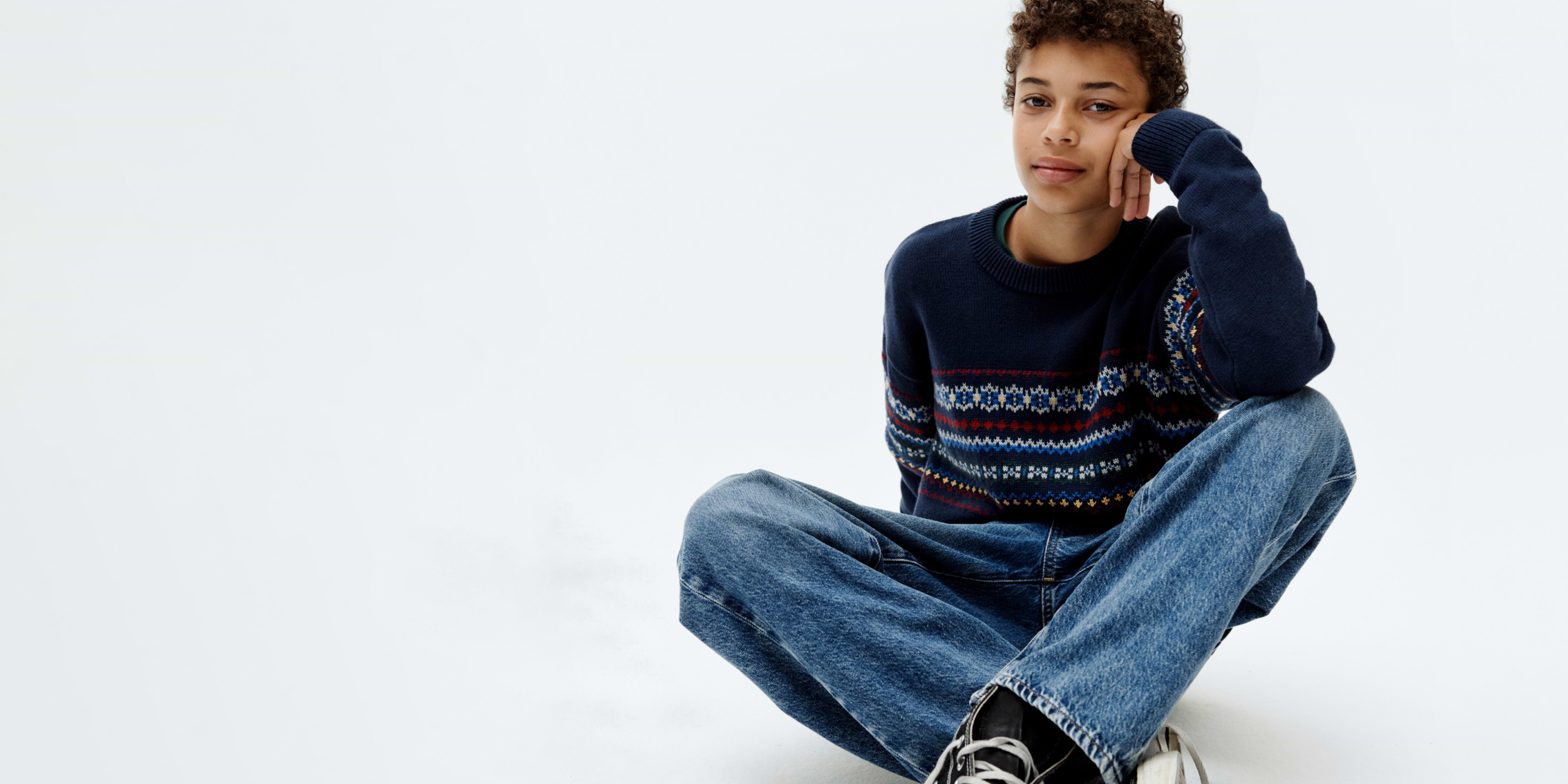 Person in a navy patterned sweater and jeans sits cross‑legged, resting chin on hand.