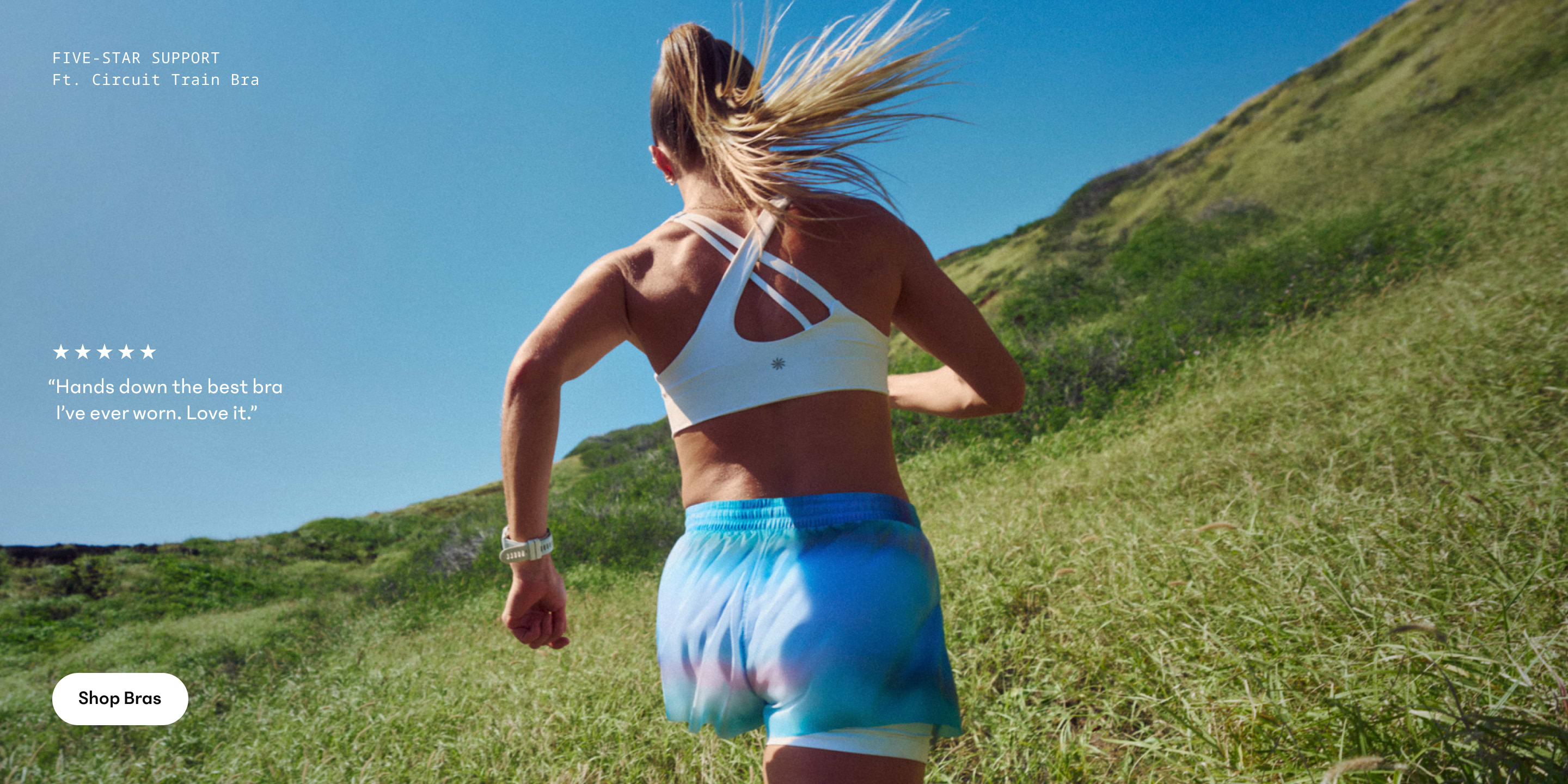 Back view of a runner in a white strappy sports bra and blue shorts sprinting up a grassy hillside under a clear blue sky.