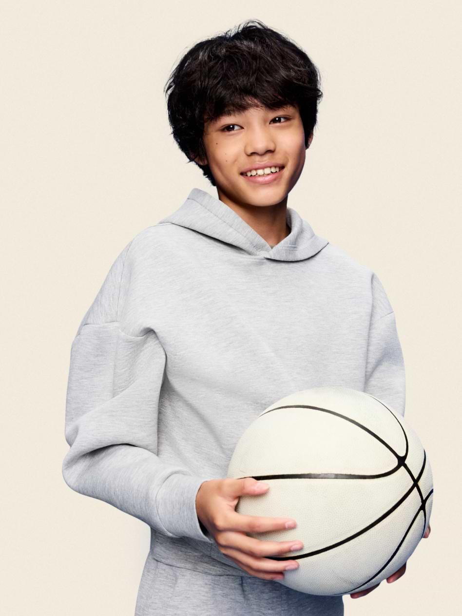 Smiling young person in a gray hoodie holding a white basketball against a plain background.