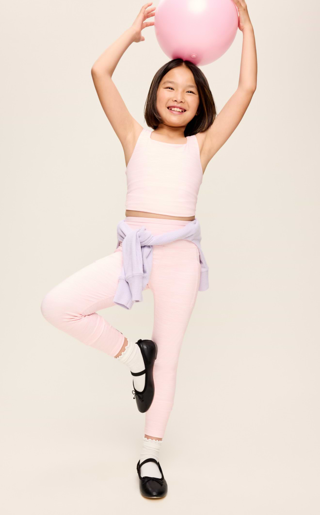 Smiling child in pastel pink activewear balances on one leg, holding a pink ball overhead with a light purple sweater tied at the waist.