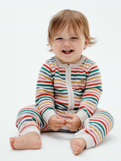 Smiling toddler sits barefoot on a white background, wearing a colorful striped zip-up onesie.