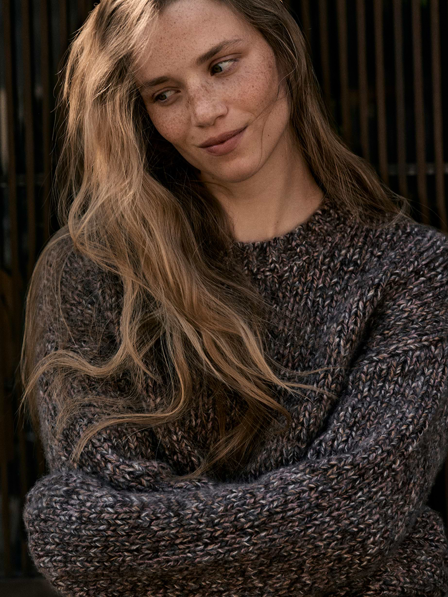 Person with long wavy hair in a chunky dark knit sweater, arms crossed, looking to the side with a soft smile.