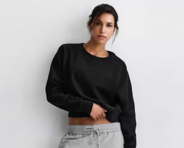 Person wearing a black crewneck sweatshirt and gray joggers, standing against a plain light wall.