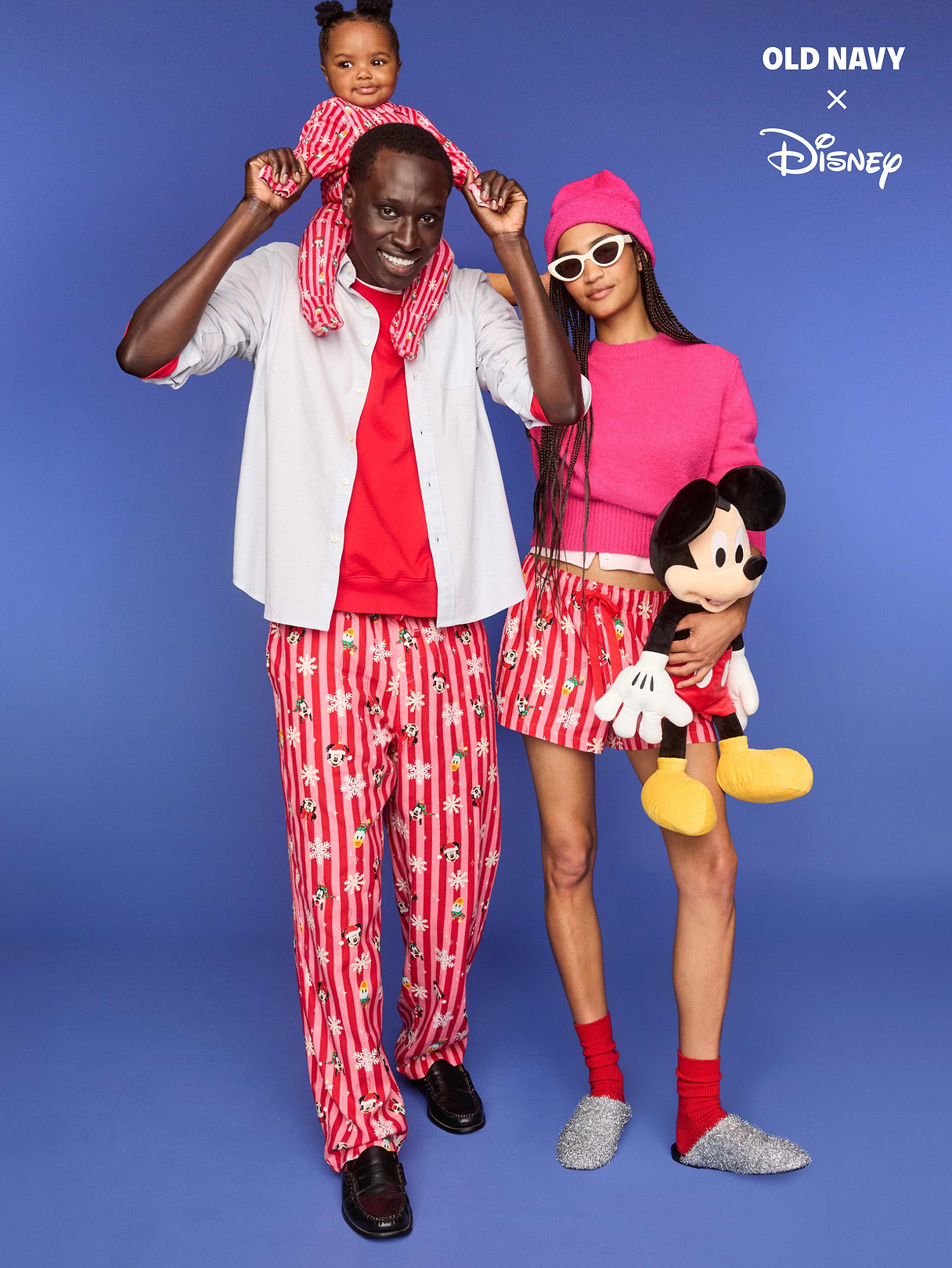 Two adults and a child in matching red Mickey Mouse pajamas; one adult carries the child on their shoulders while the other holds a Mickey plush, against a blue studio background with “Old Navy x Disney” text.