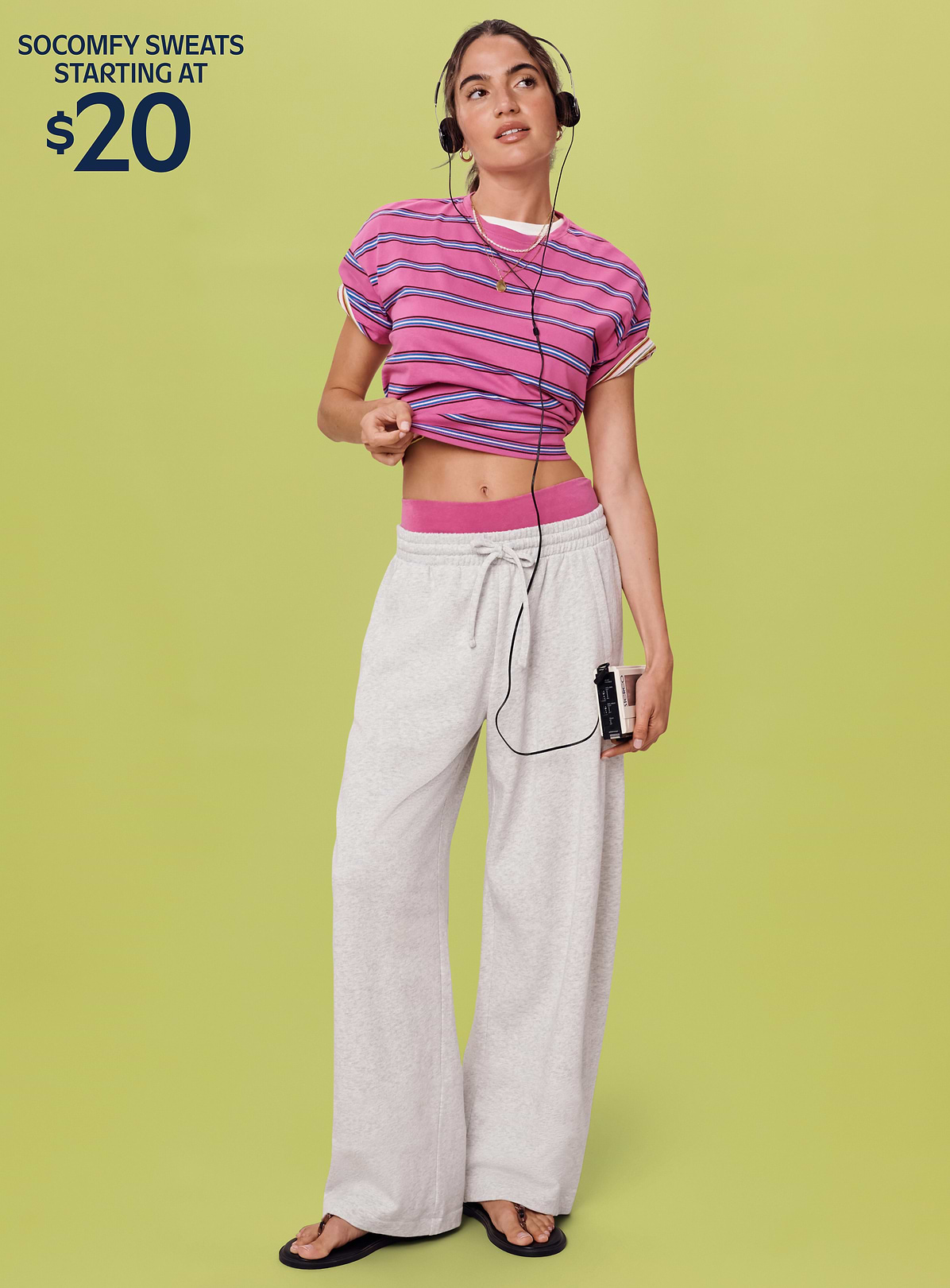 Person wearing a pink striped crop top and gray wide‑leg sweatpants, listening with headphones and holding a cassette player; green background with text “Get the look for $35.”