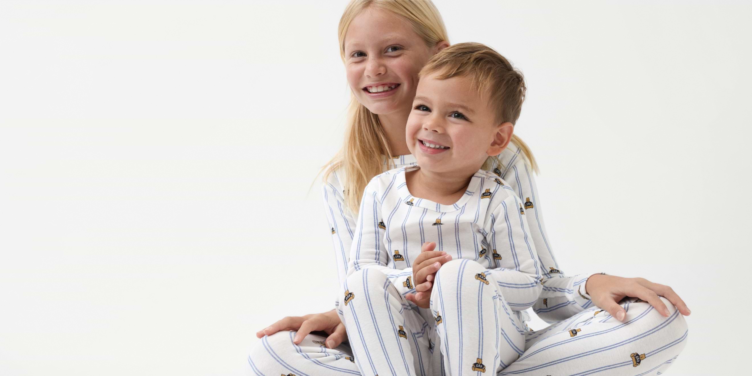 Two smiling children in matching white pajamas with blue stripes and small bear prints sit together against a white background.