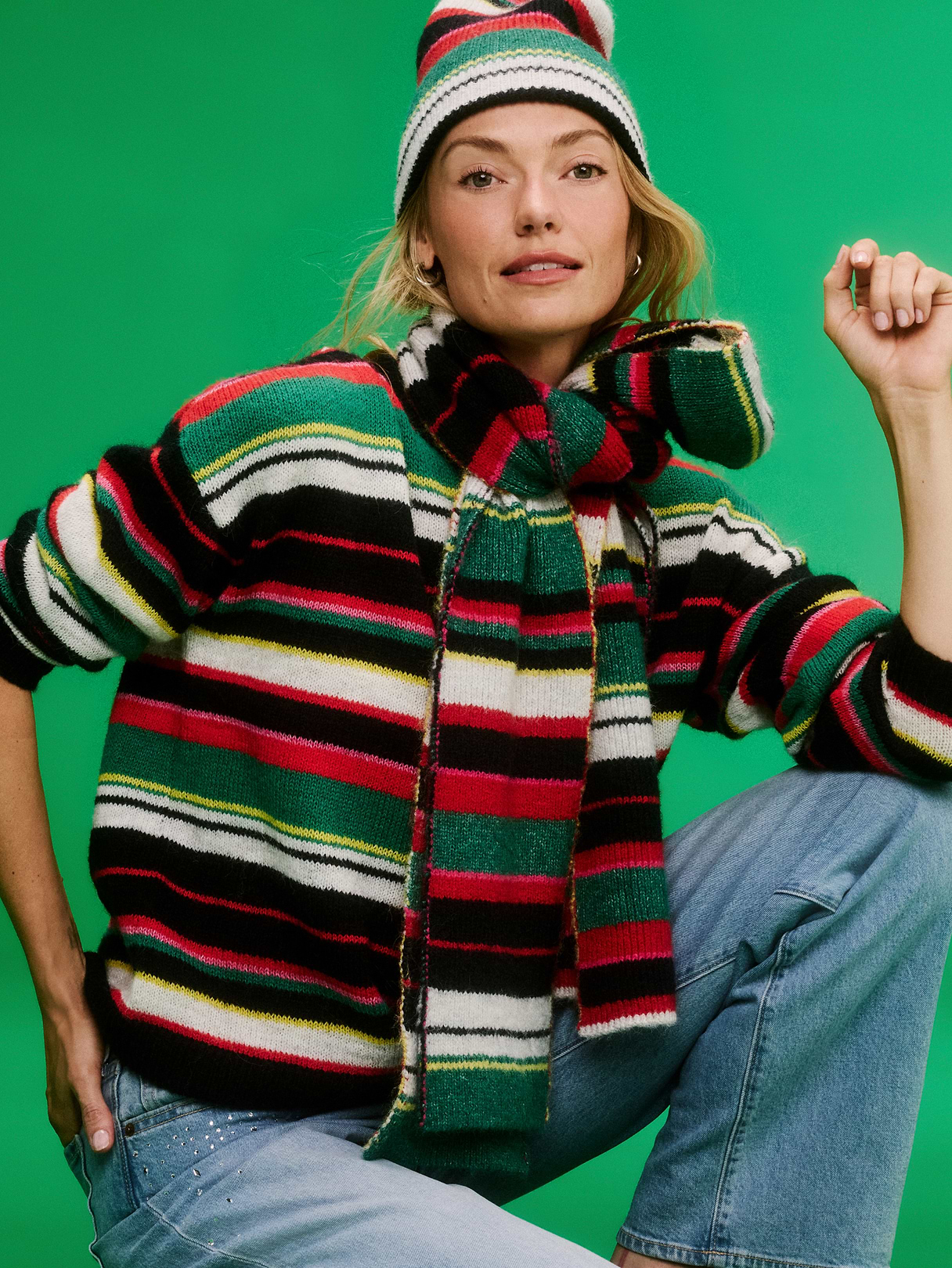 Person in a red, green, black, and white striped sweater with matching scarf and beanie, seated in light-wash jeans against a green backdrop.