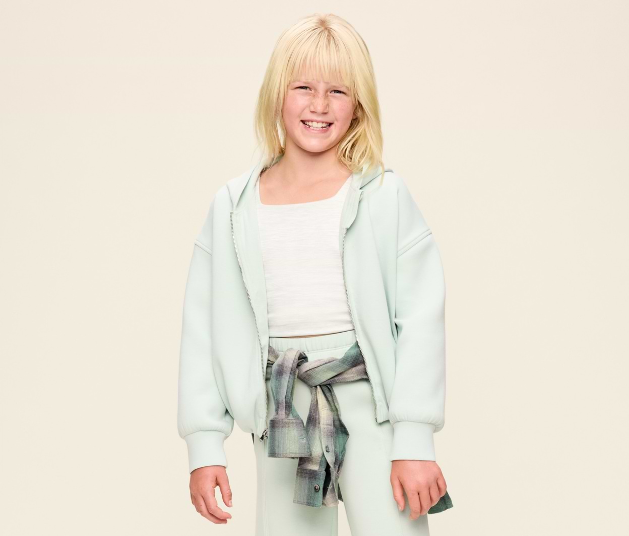 Smiling child in a mint green hoodie and sweatpants, white top, and plaid shirt tied at the waist, standing against a plain beige background.