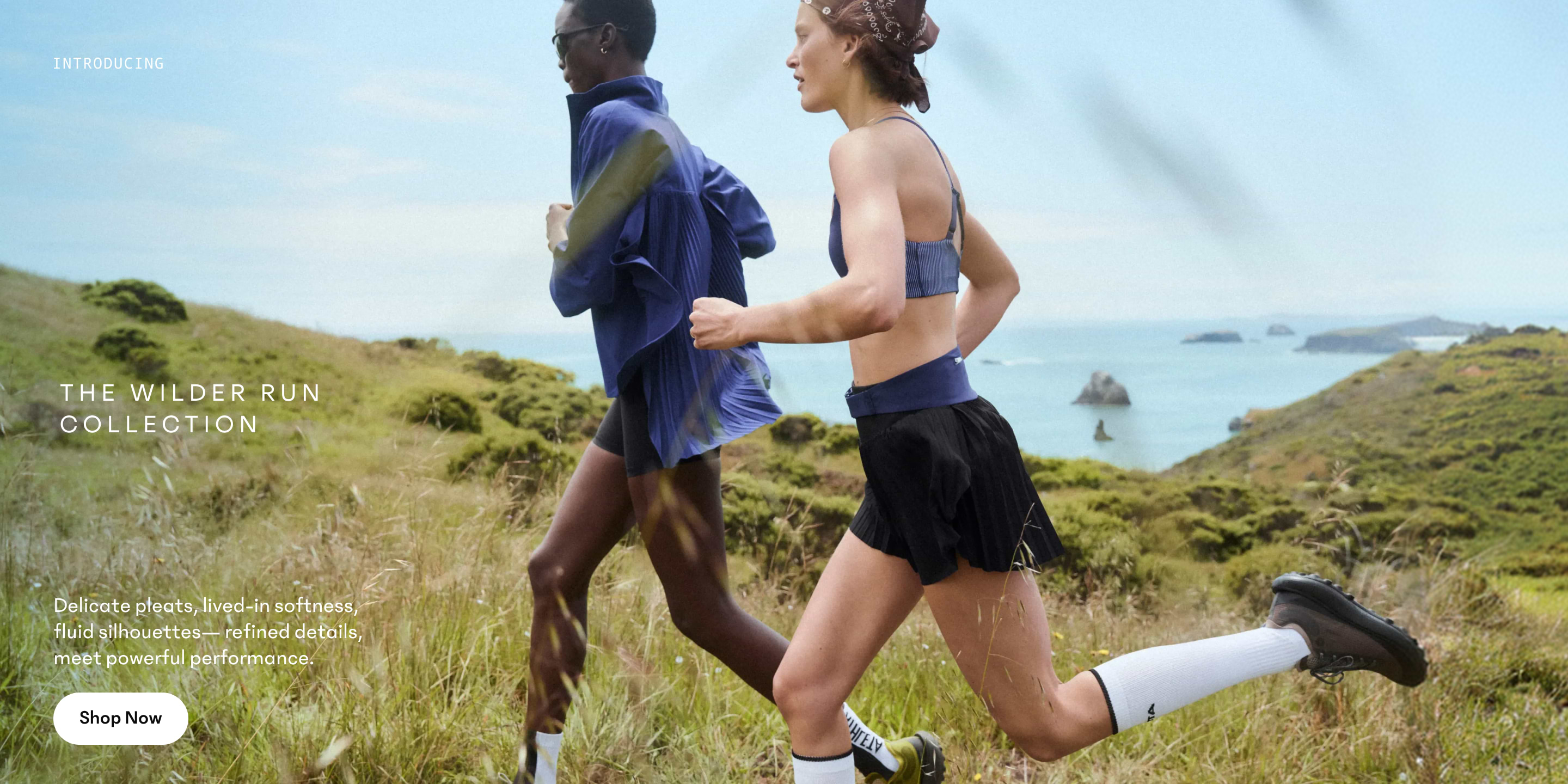 Two women running along a grassy coastal trail, wearing lightweight athletic gear with the sea and islands behind.